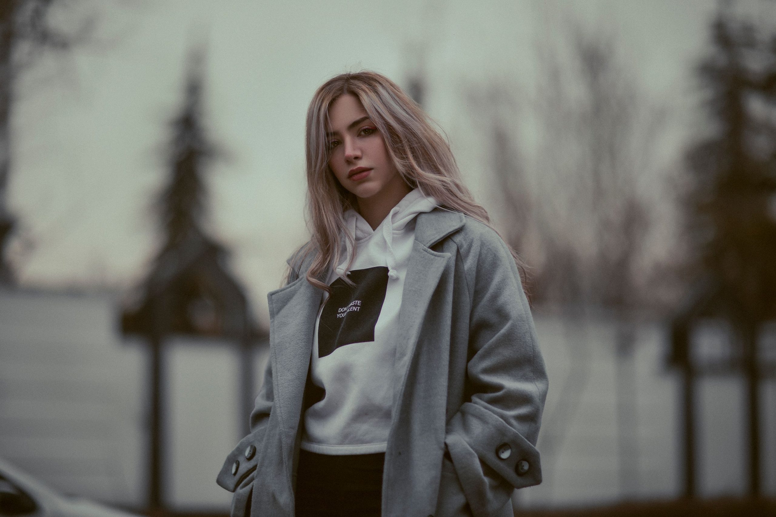 A fashionable blonde woman poses outdoors in a coat and hoodie, embodying modern winter style.