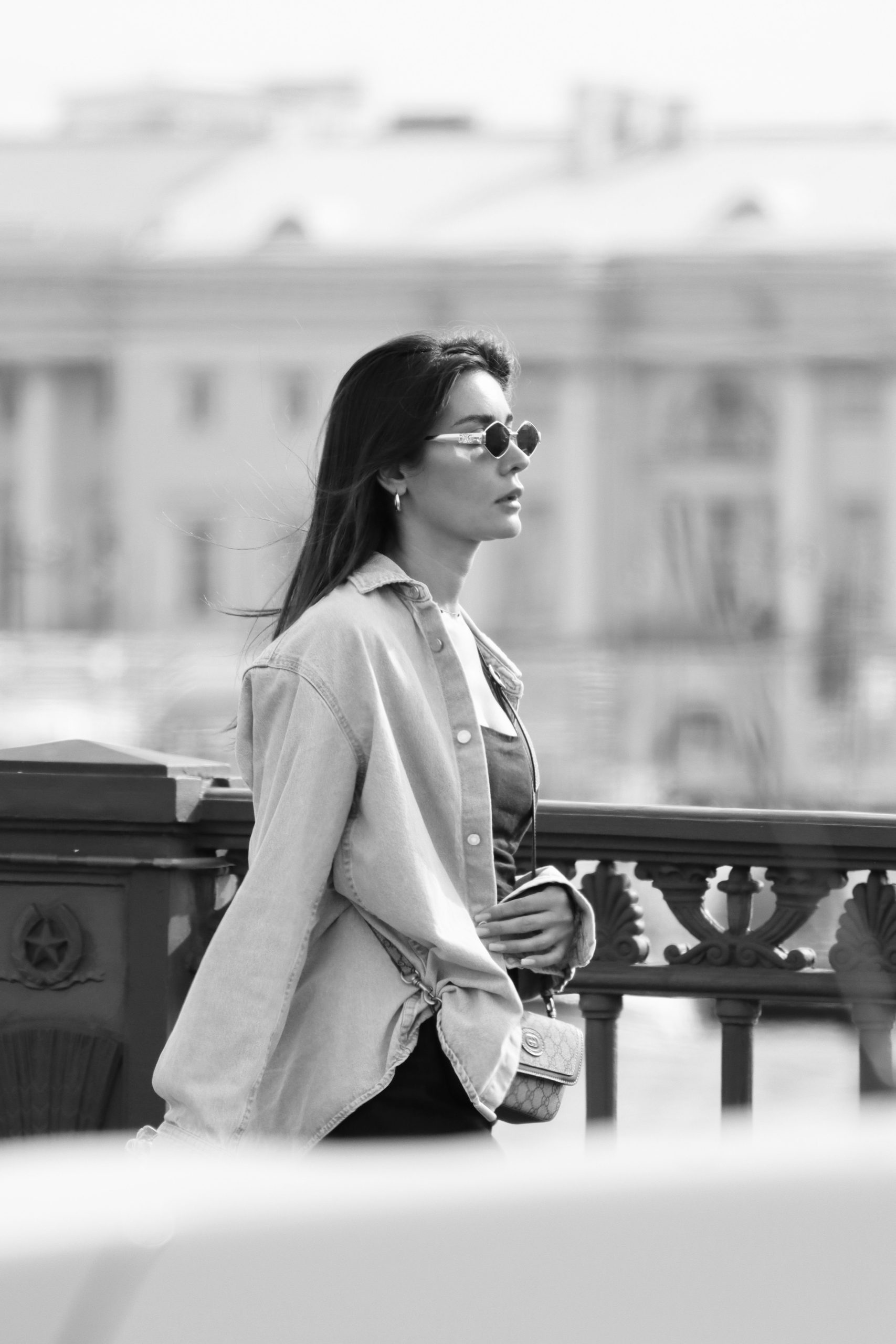 Elegant black and white photo of a fashionable woman walking near an ornate iron fence in a city setting.