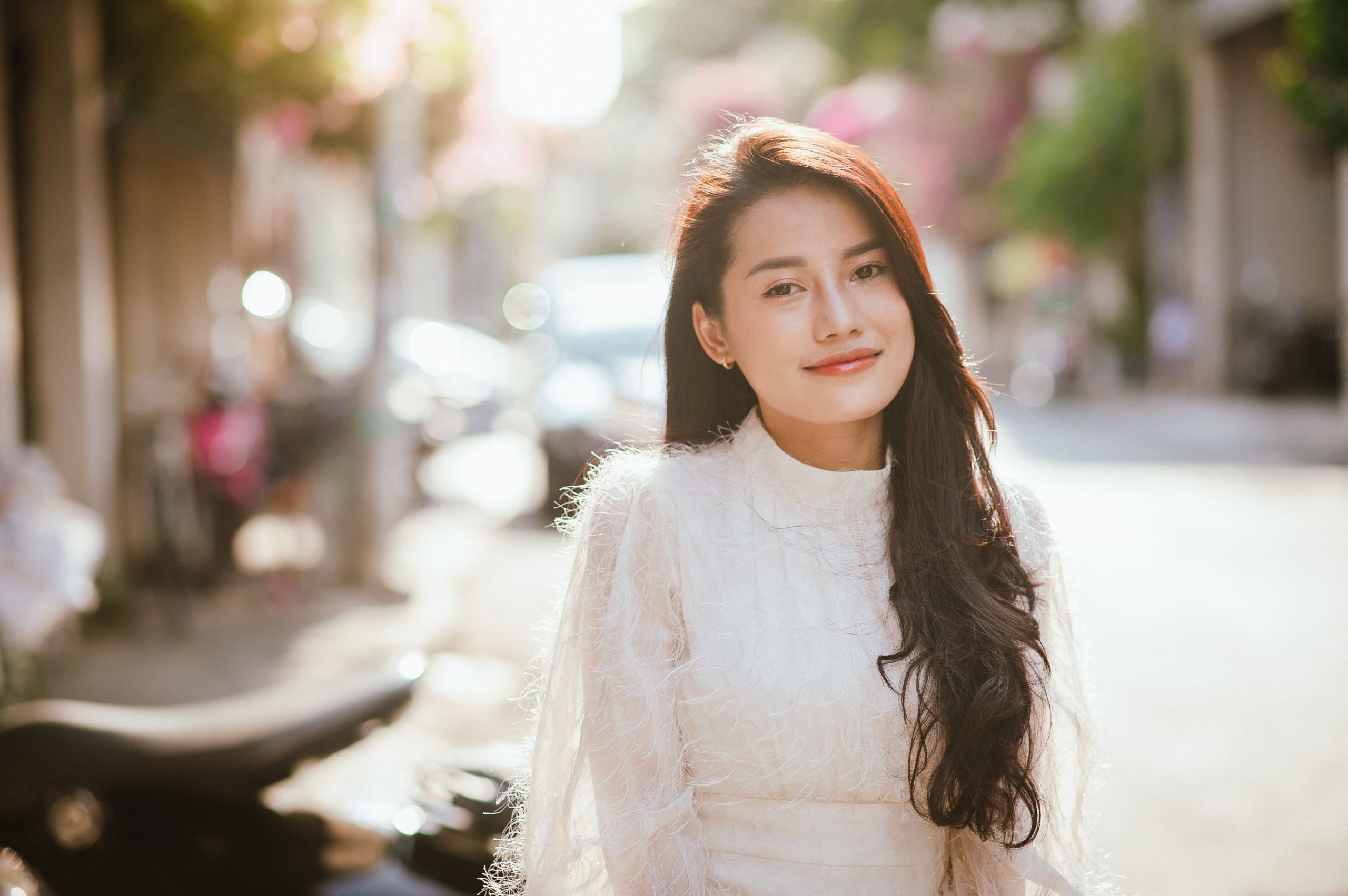 A woman in a white dress smiles warmly in a sunny street in Ho Chi Minh City.