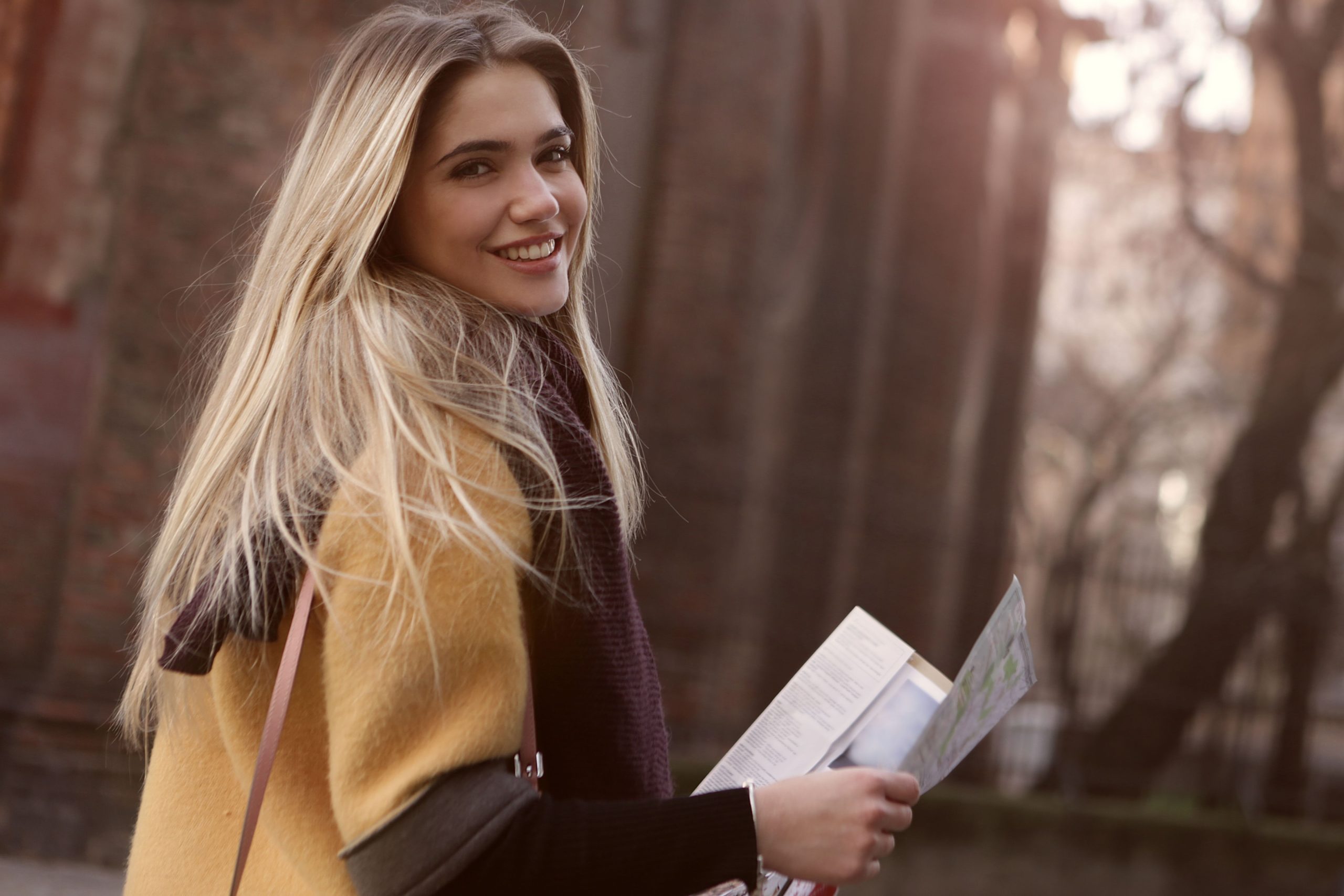 Young blonde woman smiling outdoors, holding a map on a sunny day, enjoying a casual outing.