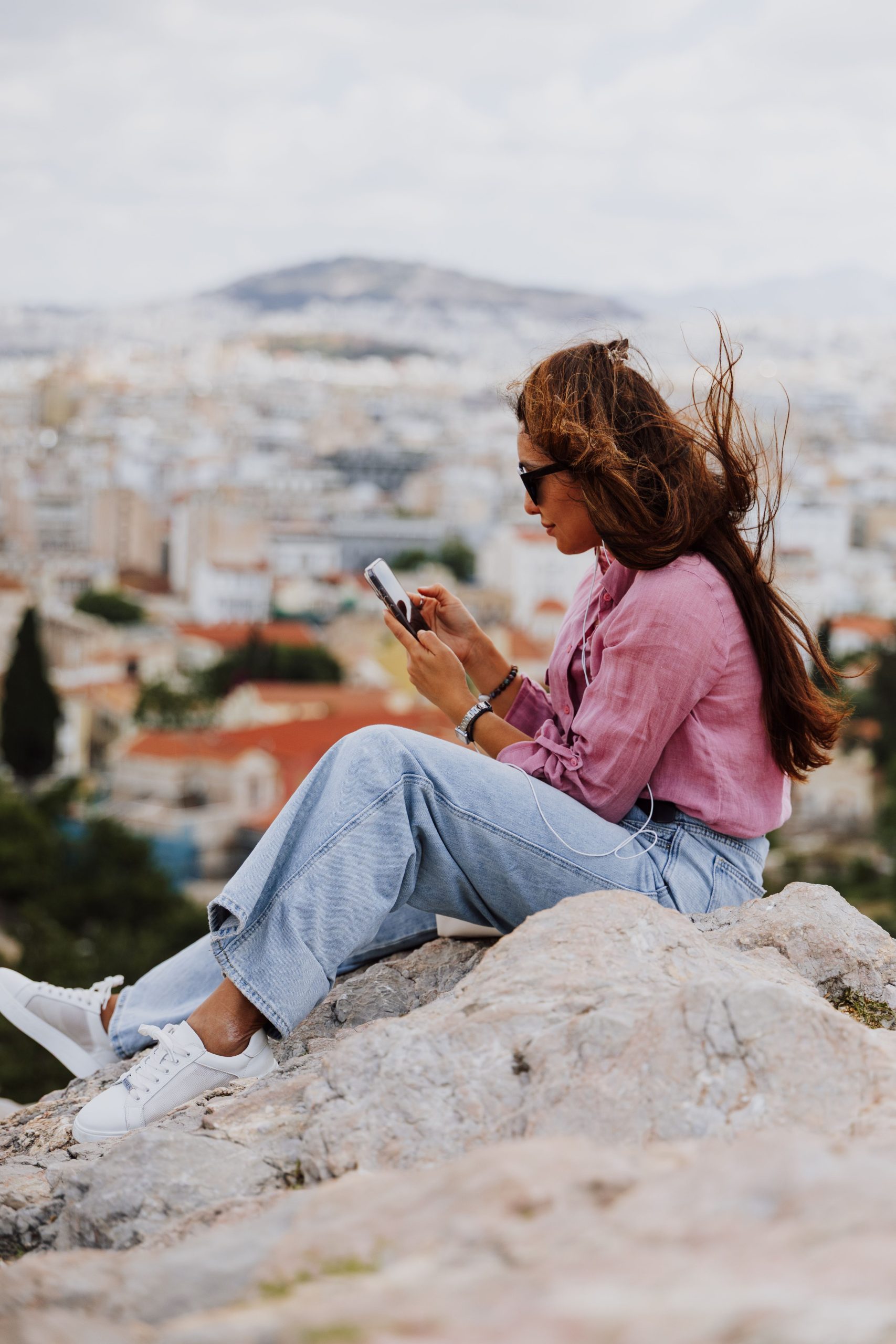 Young woman sitting on a hill using smartphone, overlooking Athens, Greece.