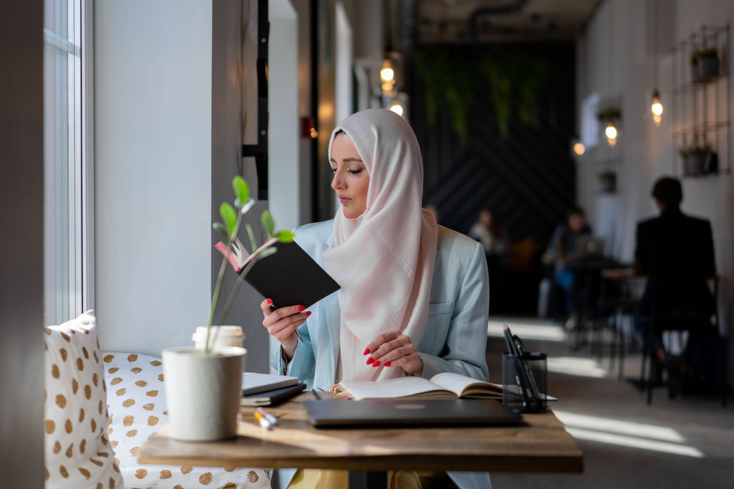 Middle Eastern woman in a hijab reading a book at a stylish cafe table.