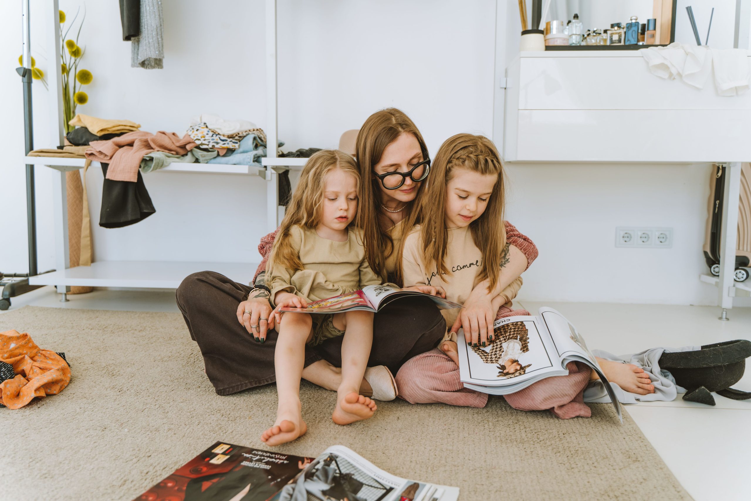 A mother and her two daughters sit on a rug indoors, enjoying magazines together.
