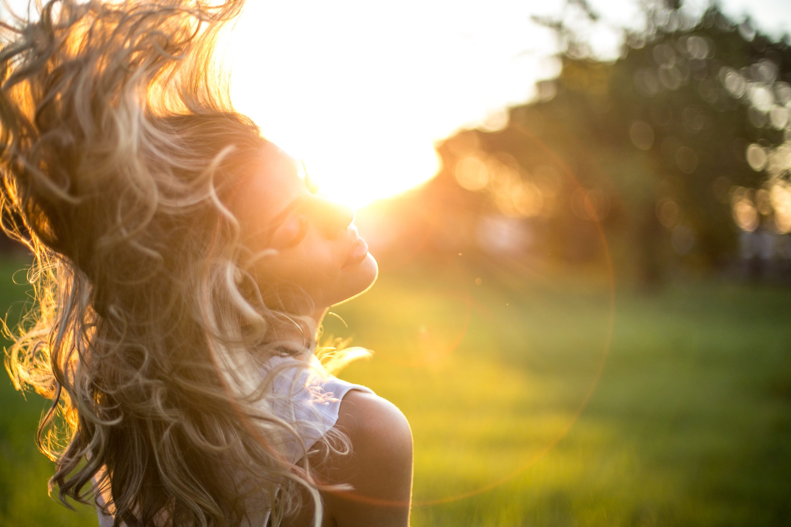 A young woman tosses her long, wavy blond hair in the sunlight during golden hour outdoors.