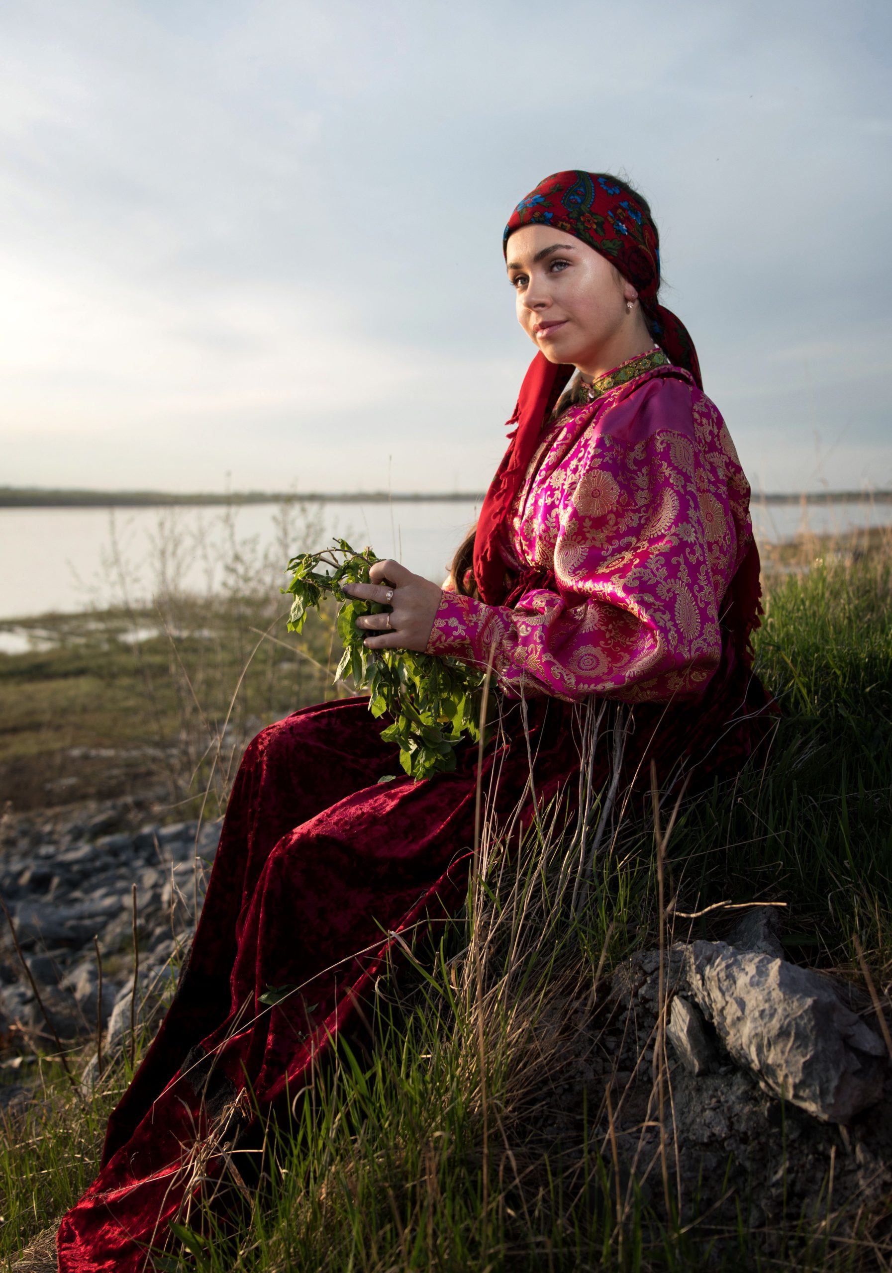 Woman in traditional clothing holding greens by a riverbank, surrounded by nature.